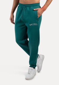 Pantalones jogger color teal con cintura elástica y cordón, que cuentan con bolsillos laterales y un logotipo bordado en blanco. Combinados con zapatillas blancas.