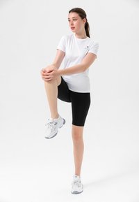 Hvit trenings t-skjorte kombinert med svarte shorts, med en tettsittende design. Bært sammen med hvite og grå treningssko. Enkelt bakgrunn.