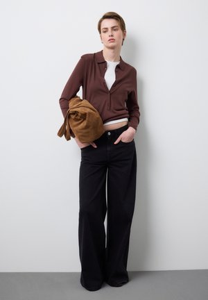 Junge Person steht vor einer weißen Wand und trägt ein braunes, geknöpftes Hemd über einem weißen Oberteil, weit geschnittene schwarze Hosen und hält eine braune Wildledertasche.