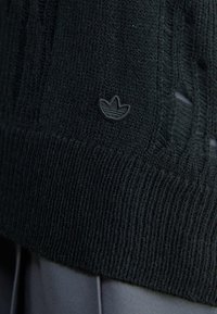 Svart stickad tröja med ribbad textur och en liten broderad logotyp. Den nedre fållen visar en blandning av täta och lösa stickade mönster.