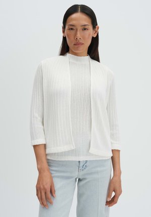 Femme aux cheveux bruns lisses portant un cardigan blanc côtelé sur un haut blanc assorti et un jean bleu clair, debout devant un fond uni.