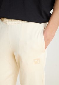 Cream-colored sweatpants with an elastic waistband, featuring a small white Puma logo patch on the left side and side pockets.