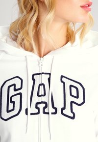Mujer rubia vestida con una sudadera blanca de GAP con cremallera y grandes letras en azul contorneadas, mirando hacia la derecha con los labios ligeramente entreabiertos.