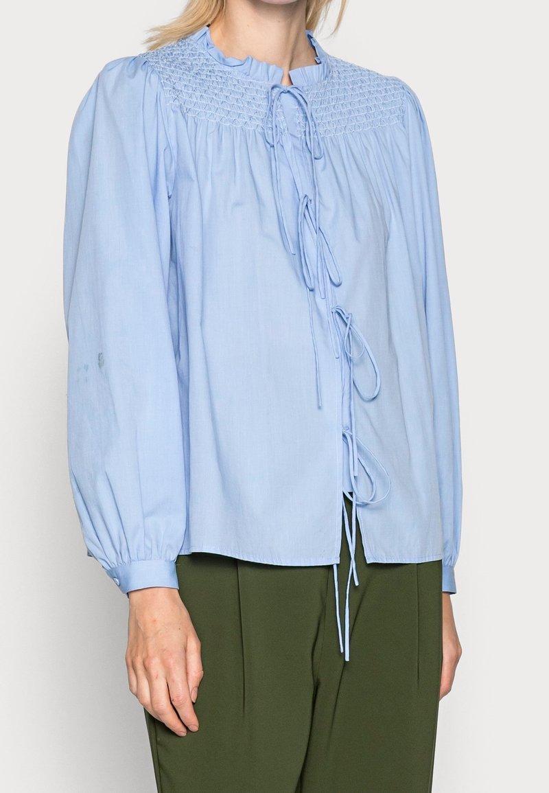 Femme portant un chemisier bleu clair à manches longues avec un empiècement froncé et des liens sur le devant, associé à un pantalon vert olive.