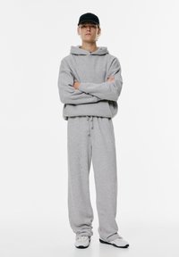Sivá bombažna kapuca z vrvico, v paru z ohlapnimi trenirkami v isti barvi. Outfit dopolnjuje črna kapa in bele teniske.