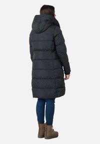 Donna che indossa un lungo cappotto imbottito nero con cappuccio, jeans blu e stivali marroni con lacci, in piedi girata di spalle su uno sfondo bianco.