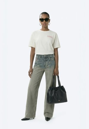 Žena v bielom tričku Diesel, sivé široké rifle s vysokým pásom a vyblednutým vzhľadom, čiernymi špicatými topánkami, okuliarmi cat-eye a drží čiernu koženú tašku s nariasením.