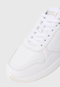 Λευκά δερμάτινα sneakers με λευκά κορδόνια και χρυσό λογότυπο "Tommy Hilfiger" στη γλώσσα του παπουτσιού, δείχνοντας λεπτομερή ραφή στο πάνω μέρος.