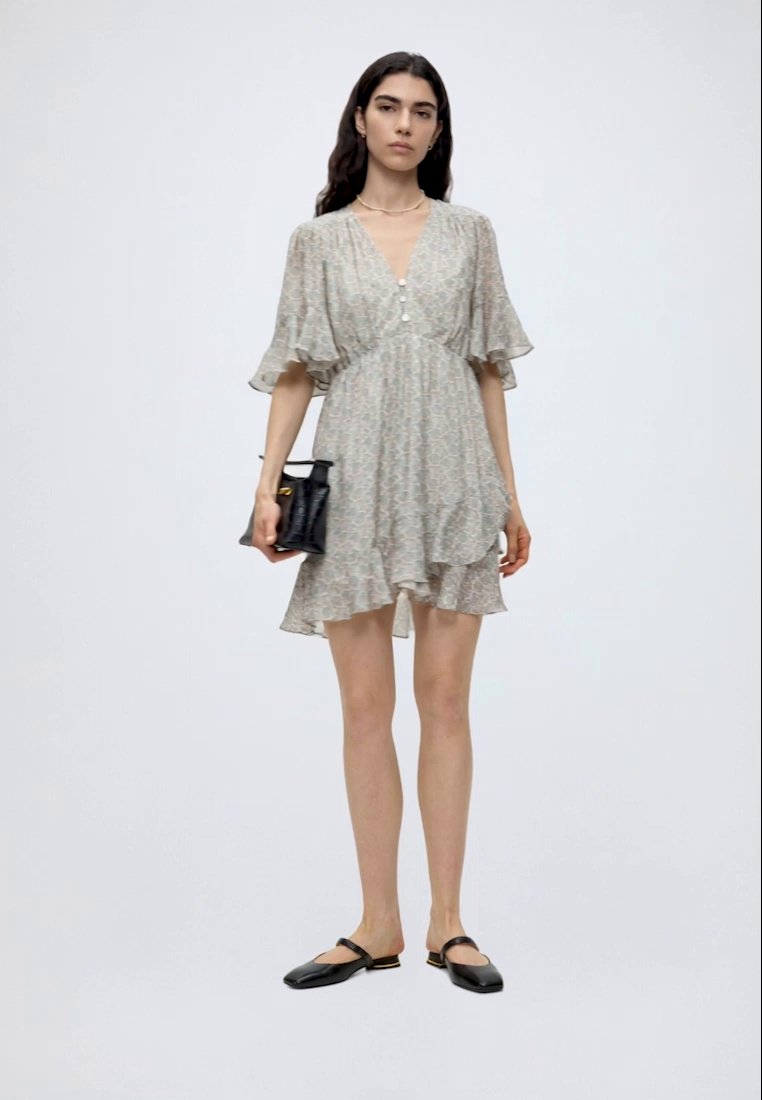 Femme portant une robe courte gris clair à motifs avec des manches volantées, tenant un sac à main noir et portant des chaussures plates noires.