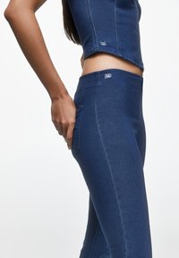 Kvinna i mörkblå figurnära denimleggings och matchande ärmlös topp, med handen vilande på sidofickan mot en vit bakgrund.