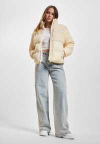 Puffig beige jacka med en texturerad fleecepanel vid bröstet, med dragkedjestängning och broderad branding framtill.