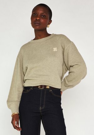 Donna che indossa un maglione beige a maniche lunghe con un piccolo logo, jeans scuri a vita alta, orecchini a cerchio dorati e braccialetto, in piedi con una mano sul fianco.