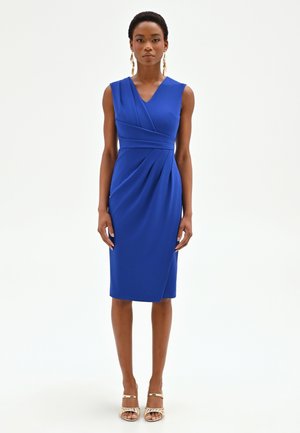 Femme portant une robe portefeuille bleu royal sans manches, longueur genou, avec des détails plissés, des boucles d'oreilles pendantes dorées et des talons ouverts dorés, debout contre un fond blanc.