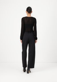 Long-sleeve black mesh top with ribbed texture, paired with high-waisted black trousers, seen from the back, with block-heeled shoes.