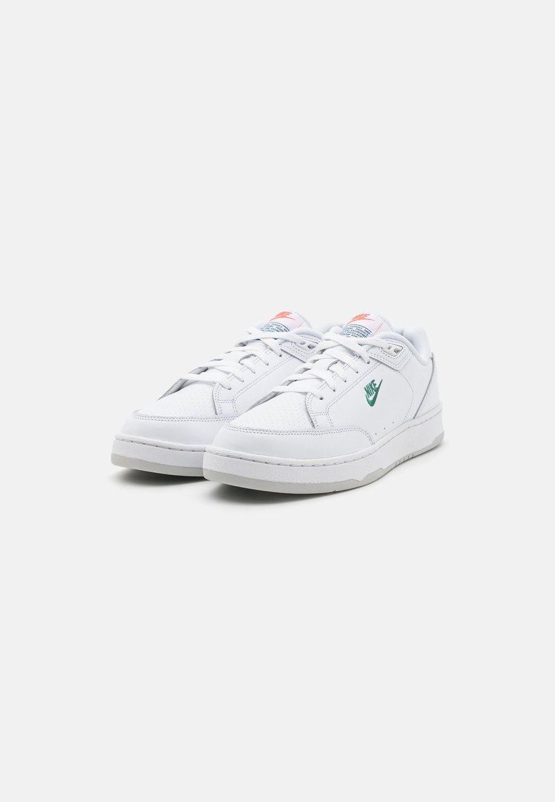 Baskets blanches avec une tige en cuir texturé, logo Nike vert et accents roses sur la languette. Lacets plats et semelle en caoutchouc.