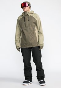 Snow jacket in olive green and beige with hood, front pocket, and adjustable drawstrings. Paired with black snow pants and black shoes.