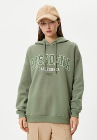Sudadera verde oliva hecha de tela suave con capucha ajustable. Presenta el texto en blanco y verde "PASADENA CALIFORNIA 1991" en el frente.