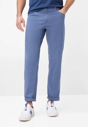 Mand iført lyseblå afslappede bukser med opkrogede manchetter, hvide sneakers med blå accenter og en mørkeblå skjorte, stående på hvid baggrund.