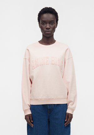 Femme portant un sweat-shirt oversize rose clair avec des lettres texturées "ANINE BING" et un jean bleu large, debout devant un fond blanc uni.