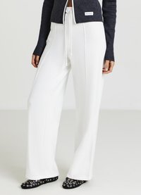 Pantalon blanc à jambes larges avec une texture lisse, taille à cordon de serrage, et coutures latérales contrastantes, associé à des chaussures noires ornées d'accents argentés.