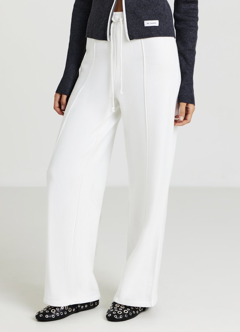 Pantalon blanc à jambes larges avec une texture lisse, taille à cordon de serrage, et coutures latérales contrastantes, associé à des chaussures noires ornées d'accents argentés.