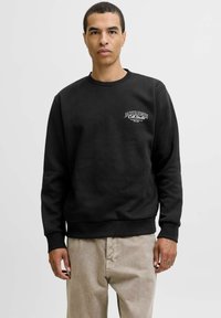 Mand iført sort sweatshirt med "Jack & Jones Col. Studio" logo og beige bukser, stående foran en ensfarvet lys baggrund.