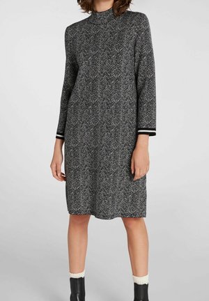 Kobieta ubrana w czarno-białą sukienkę o długości do kolan w jodełkę, z długimi rękawami, czarne botki za kostkę oraz białe skarpetki.