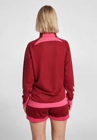 Chaqueta deportiva burdeos con acentos rosas y detalles de malla, combinada con pantalones cortos burdeos a juego con ribetes rosas.