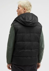 Schwarze Pufferweste mit Kapuze, die über gesteppten Bereichen und einer matten Oberfläche verfügt. Getragen über einem strukturierten grünen Langarmshirt.