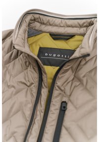 Beige gesteppte Jacke mit schwarzem Reißverschluss und gelber Fütterung. Das Etikett trägt die Aufschrift "bugatti", mit einer angehängten Stoffschlaufe am Kragen. Strukturmuster.