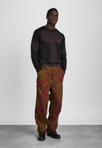 Μαύρο μακρυμάνικο μπλουζάκι με στρογγυλή λαιμόκοψη; καρό παντελόνι σε κόκκινες και πράσινες αποχρώσεις με φαρδιά πόδια και τσέπες; φορεμένο με σκούρα παπούτσια σε ουδέτερο φόντο.