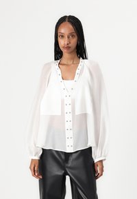 Blusa bianca trasparente con maniche a sbuffo, design con bottoni e dettagli in metallo, abbinata a pantaloni di pelle nera. Texture liscia, vestibilità comoda.