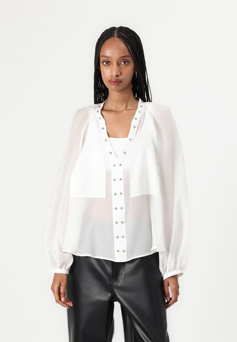 Blusa bianca trasparente con maniche a sbuffo, design con bottoni e dettagli in metallo, abbinata a pantaloni di pelle nera. Texture liscia, vestibilità comoda.