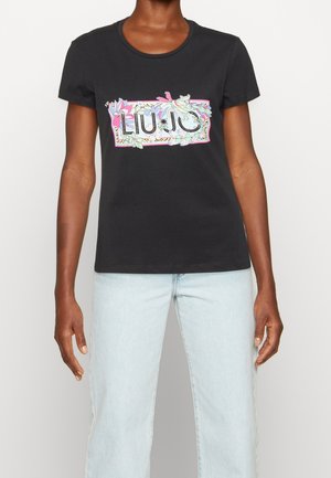 Frau trägt ein schwarzes Kurzarm-T-Shirt mit buntem floralen „LIU JO“-Logo und hellblauen Jeans vor einfarbigem Hintergrund.
