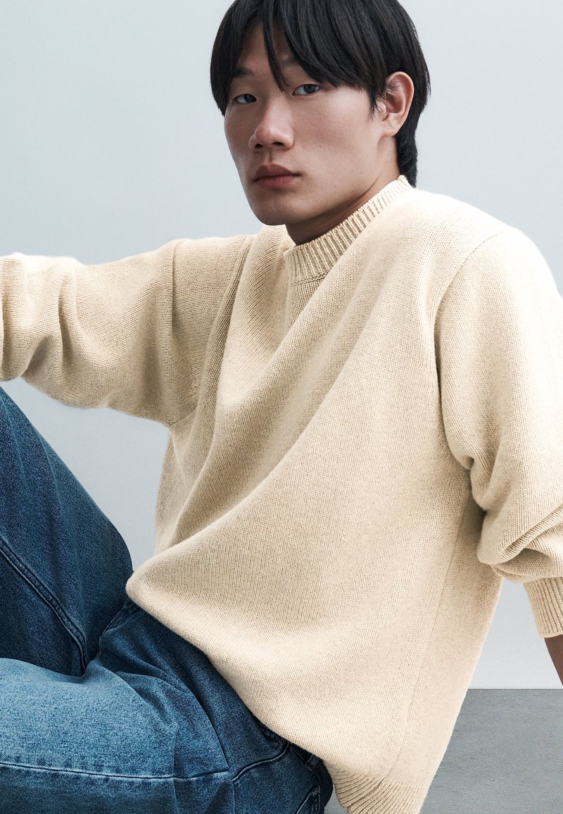 Jeune homme aux cheveux noirs assis, portant un pull en tricot beige et un jean bleu, sur un fond uni gris clair.