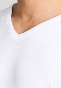 Vit bomulls T-shirt med V-ringning, med en mjuk textur och en subtil lyster. Sömmarna är synliga vid halsringningen och axeln för att ge extra detalj.