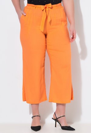 Frau trägt leuchtend orangefarbene, weit geschnittene, kniehohe Hosen mit gebundenem Taillenband und schwarzen Slingback-Pumps mit hohem Absatz, stehend vor einem weißen Hintergrund.