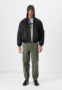 Schwarze Bomberjacke, weißes T-Shirt mit blauem und gelbem Grafikdesign, grüne Cargo-Hosen, schwarze Sneakers. Das Modell steht mit den Händen an den Seiten.