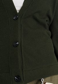Dunkelgrüner gerippter Strickcardigan mit V-Ausschnitt, schwarzen Knöpfen und einer figurbetonten Passform. Texturiertes Material und auffälliger vertikaler Rippeneffekt.
