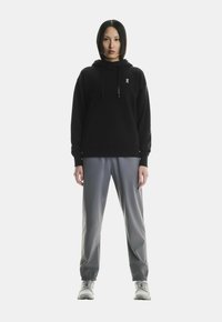 Must mustaga kapuutsiga sviiter, millel on paelad, koos hallide kitsaste pükstega. Pükstel on elastsed mansetid. Jalatsiteks on heledad hallid tossud.