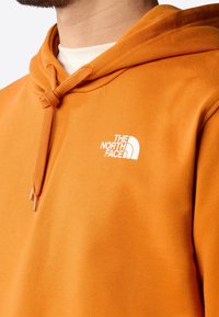 Uomo che indossa una felpa arancione con il logo bianco "The North Face" sul petto sinistro e i lacci tirati in un nodo.