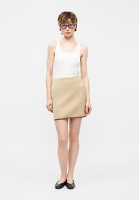 Άτομο με μπλε γυαλιά, άσπρο αμάνικο πάνω μέρος, μπεζ καρό μίνι φούστα και μαύρα παπούτσια, στέκεται μπροστά σε απλό ανοιχτό φόντο.