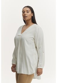 Blouse à manches longues, couleur crème, avec un col en V, en tissu texturé, fentes latérales et détails de boutons sur les manches. Matière lisse et légère.