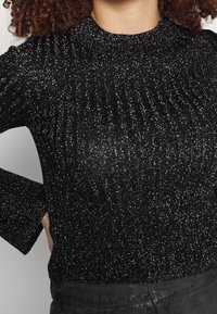 Kvinna som bär en svart glittrig ribbad långärmad tröja med hög krage, i kombination med mörka byxor, sedd från halsen till höfterna.