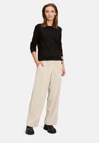 Schwarzes Langarmshirt mit rundem Halsausschnitt, kombiniert mit hellbeigen Corduroy-Hosen im weit geschnittenen Stil. Es werden schwarze Plateauschuhe getragen.