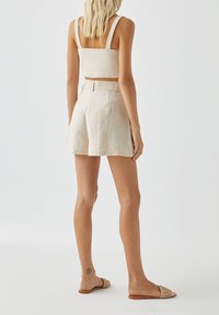 Pantaloni corti beige in lino con vita alta e gambe larghe, abbinati a un top corto beige chiaro. Il modello indossa sandali intrecciati color beige.