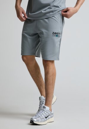 Muž nosící šedé šortky Zavetti a sladěnou košili, upravující lem košile, s bílými sportovními botami Nike na světlém pozadí.