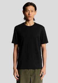 Sort bomulds T-shirt med korte ærmer, rund hals og et diskret logo på brystet. Bæres sammen med olivengrønne cargo shorts.