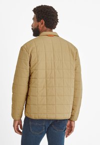 Beige quilted jacket with a straight cut and stand-up collar. Features a subtle texture and orange accent on the back. Worn with blue jeans.