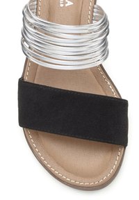 Zwarte suede instapsandalen met een brede band en een zilveren ronde band over de bovenkant. Getextureerde tan voetbed met witte stikselaccenten.